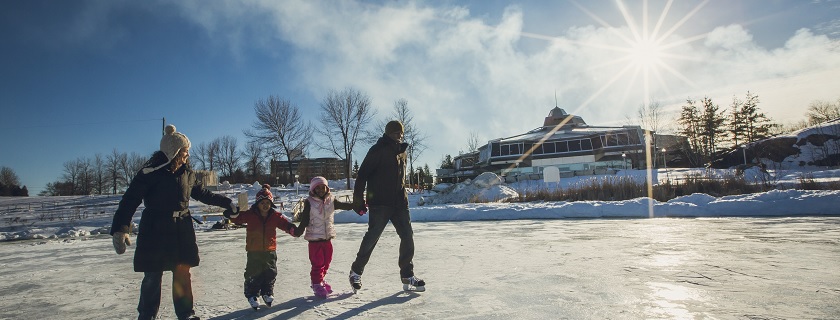 Deux adultes et deux enfants patinent sur le sentier de patinage du lac Ramsey en se tenant par la main. En arrière-plan : Science Nord.